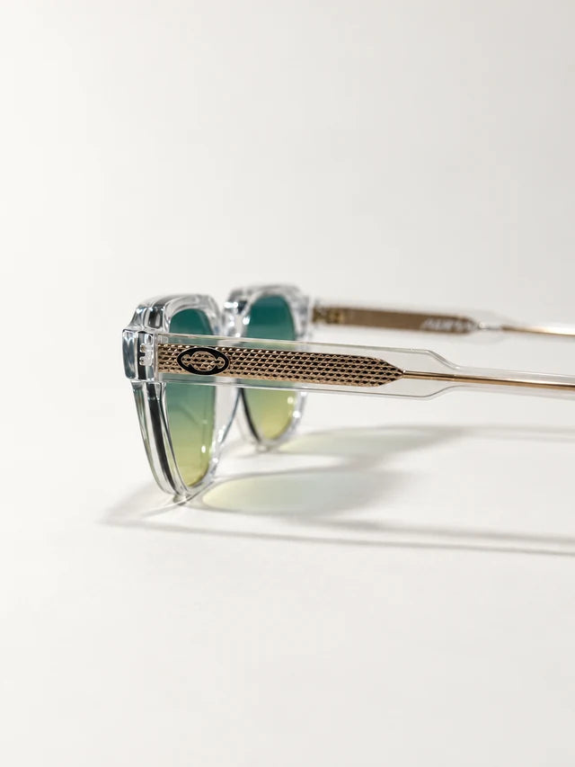 Auracle AUR-001 C5. Side detail of clear sunglasses showing a gold-tone textured temple insert and eye emblem beside green gradient lenses.
